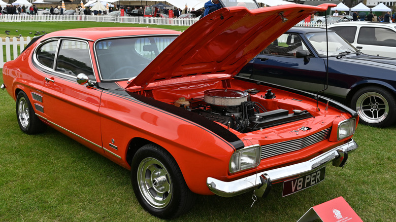 A bright orange classic Ford Capri Perana V8 coupe parked on a green lawn at an outdoor automotive exhibition.