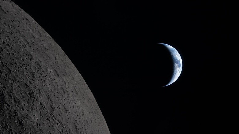 A distanct crescent Earth behind the dark cratered surface of the moon, approaching Earthset