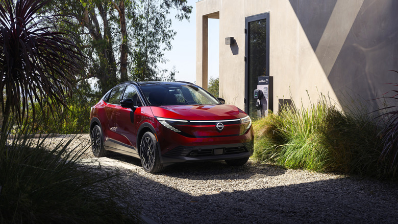 Front 3/4 view of a red 2026 Nissan Leaf parked in a driveway next to an EV charger