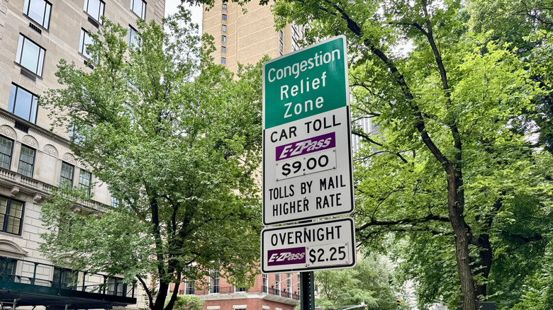 Cars driving down Fifth Avenue next to traffic cones creating temporary "Match Day" bus lane during FIFA World Cup and "Congestion relief zone: Car toll E-ZPass $9, Tolls by mail higher rate" and "Overnight E-ZPass $2.25" signs on the Upper East Side of Manhattan, New York City