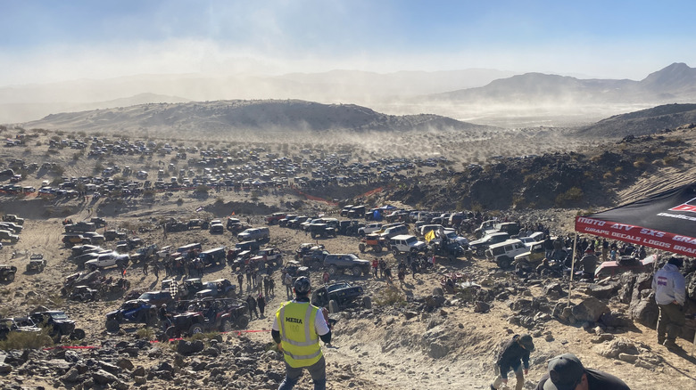 A photo from a mountain in the desert showing thousands of cars parked on the desert floor and a large dust cloud hovering above it