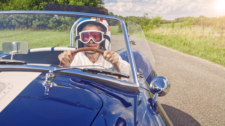 A senior female driver wearing a racing helmet and goggles speeds through rural roads in a classic Shelby Cobra.