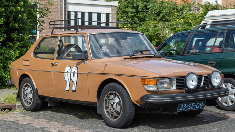 A tan Saab 99 with rally lights, a roof rack, and the number 99 painted on the side sits in a parking lot next a green station wagon and some bushes