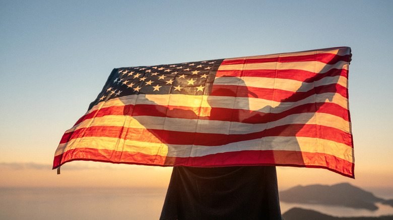 Proud man raising the flag of the United States of America standing on the top of the mountain at sunset