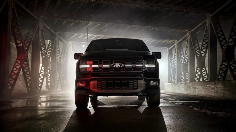 A Ford F-150's grille faintly lit, but a light bar is illuminated across the middle through the blue oval logo. The shadowed truck is backlit through the industrial bridge structure it is under.