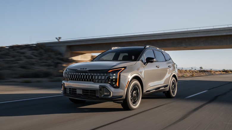 A greyish-beige Kia Sportage crossover just emerges from under a bridge on a desert highway.