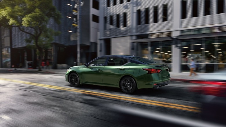 A green Nissan Altima with a black roof driving down a metropolitan city street.