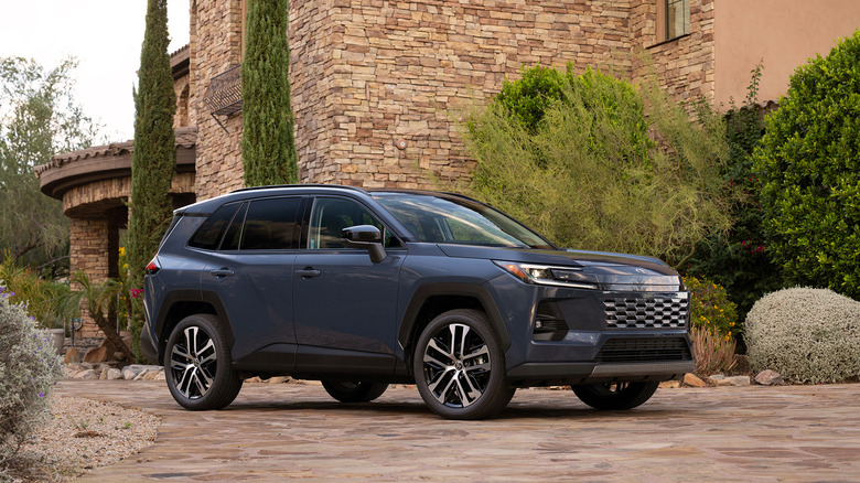 A blue-grey 2026 Toyota Rav 4 parked on a stone driveway with an older stone-built home in the background.