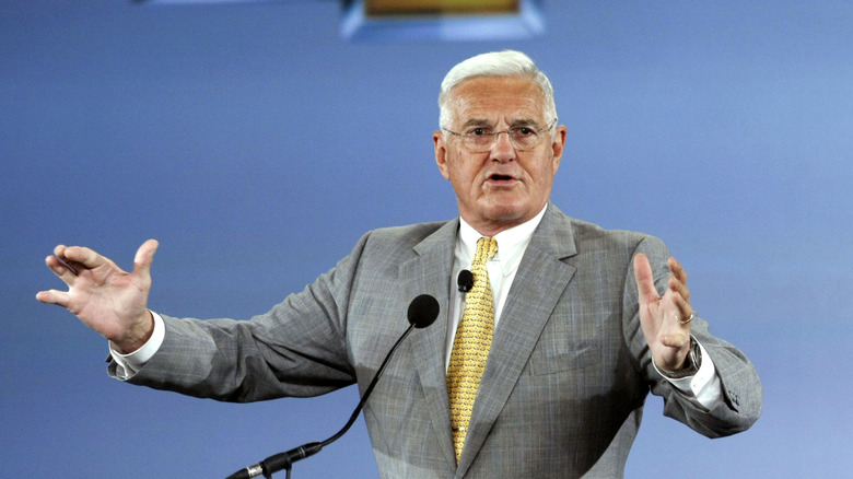Bob Lutz behind a podium and microphone, gesturing with both hands while he speaks