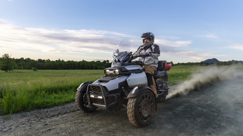 Front 3/4 view of a gray 2027 Can-Am Canyon on a dirt road next to a grassy field