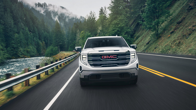 Front view of a white 2024 GMC Sierra 1500 driving through a mountainous forest next to a river
