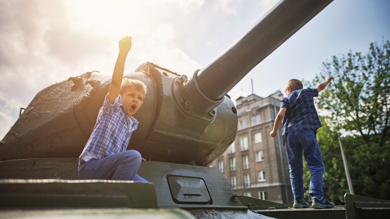 A couple of kids play on a military tank with a building in the background