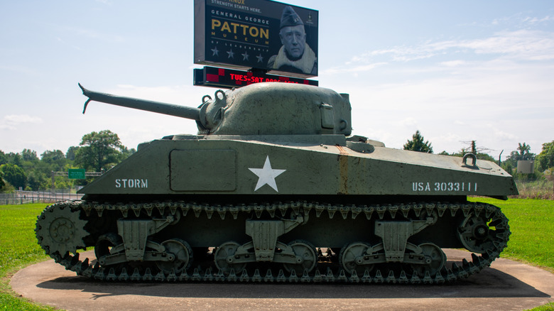 A green Sherman tank with a broken cannon barrel sits on a concrete slab in front of a billboard for the General George Patton Museum