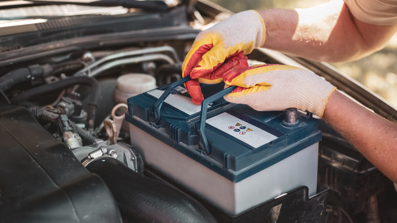 A person wearing gloves removing a car's battery from the engine bay.