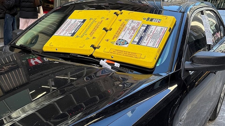 A bright yellow parking barnacle attached to the windshield of a sedan in New York City