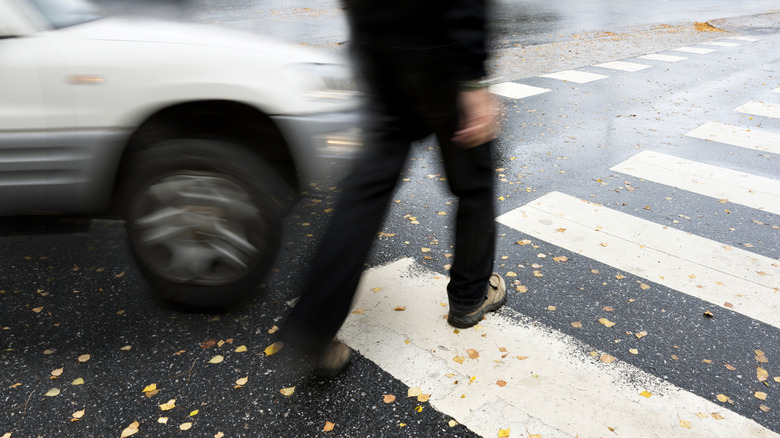 A blurred car close in proximity to the lower half of a human walking in a pedestrian crossing at an intersection.