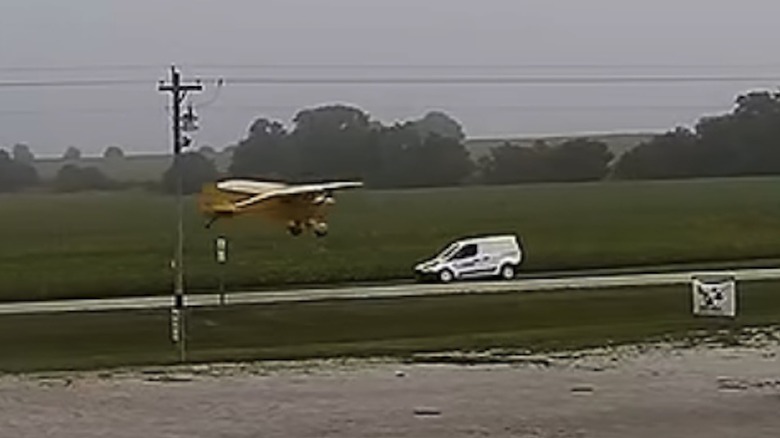 A small yellow airplane about to fly over a postal van on a narrow road between fields
