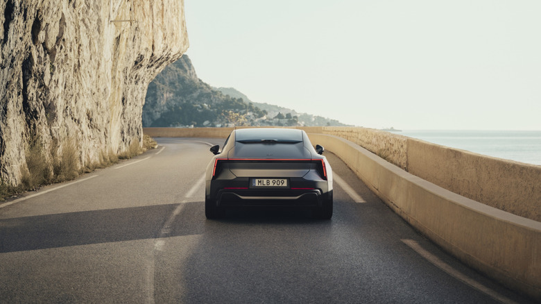 Rear view of a Polestar 5 driving down a road with a wall and water on the right and a rock cliff on the left