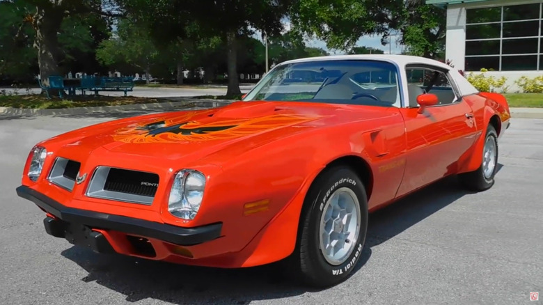 A red 1974 Pontiac Firebird Trans Am with a white vinyl Cordova roof in a parking lot in front of trees and picnic bench