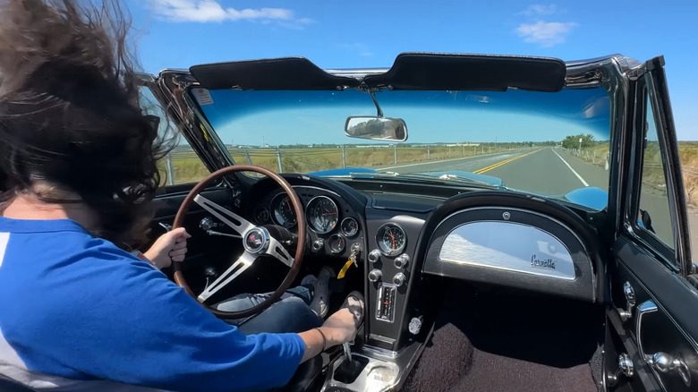 An L71 427-powered 1967 Chevrolet Corvette convertible with a four speed manual transmission being driven on a deserted road by a woman in a blue shirt with wind-blown hair