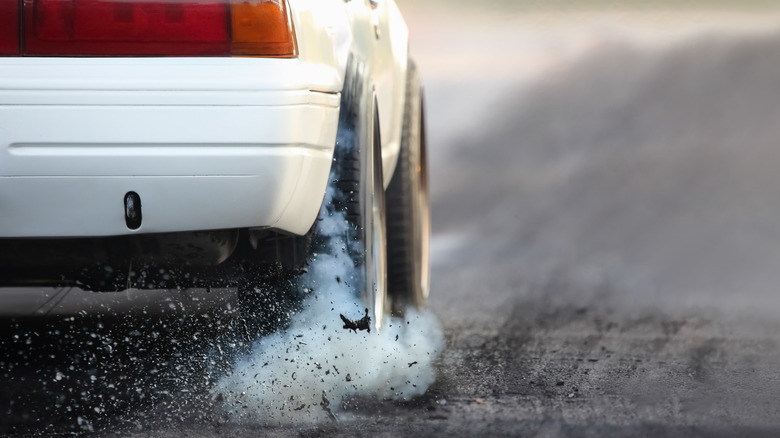 A white car doing a burnout while kicking up bits of asphalt and tire rubber