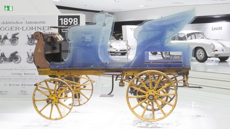 The skeletal wooden chassis of the 1898 Porsche P1 Egger-Lohner C.2 electric vehicle on display at the Porsche Museum, fitted with a translucent blue plastic body silhouette.