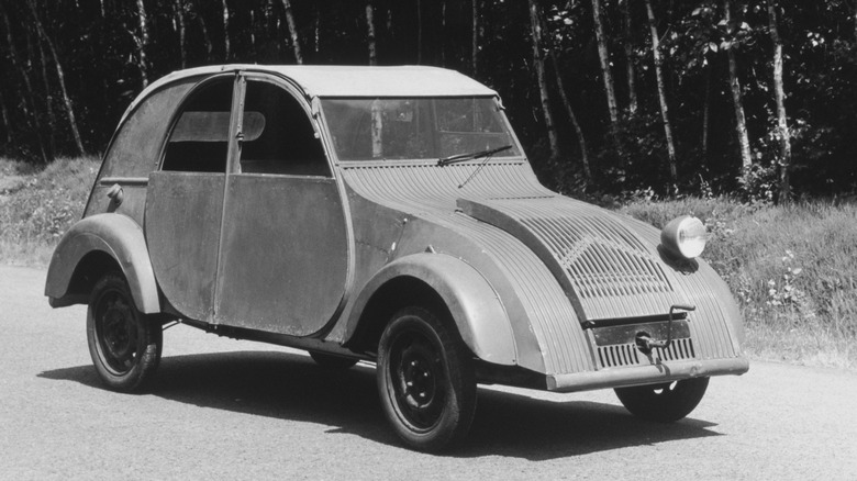 A black-and-white photograph of a minimalist 1939 Citroën 2CV TPV prototype parked on an asphalt road. You can see its signature single "cyclops" headlight and corrugated metal hood with a front hand-crank starter.