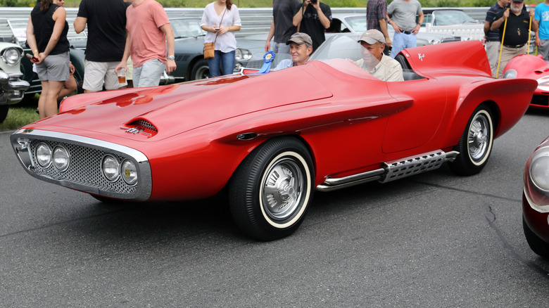 A bright red 1960 Plymouth XNR asymmetrical concept roadster driving past a crowd at an outdoor automotive event.