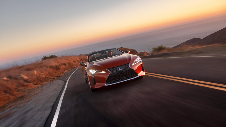 A head-on shot of a red LX500 driving up a canyon road at dusk in front of a view of the ocean
