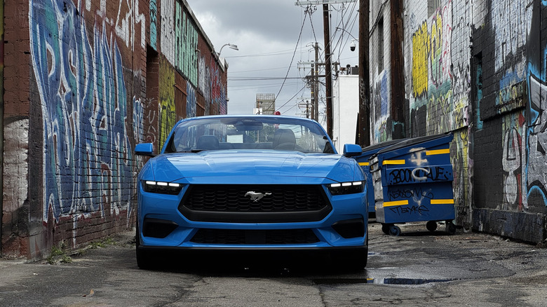 A head-on shot of a blue Ford Mustang convertible with its top down in an alley flanked by graffiti-covered brick walls