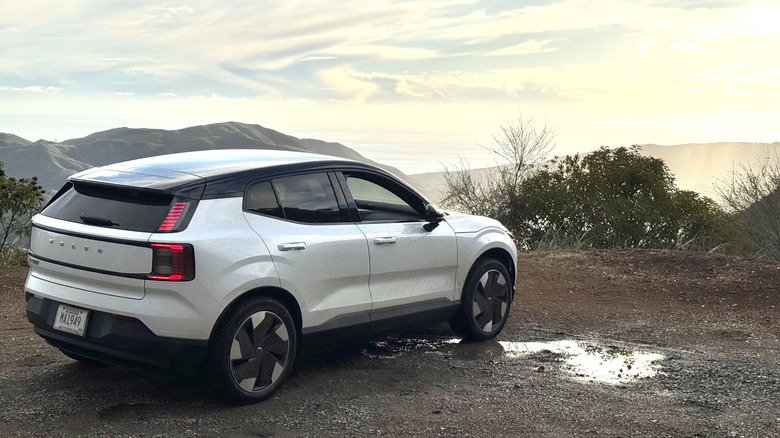 A rear three-quarters shot of the white EX30 parked on dirt next to a puddle and in front of an aerial view of mountains, the ocean, and the sunset