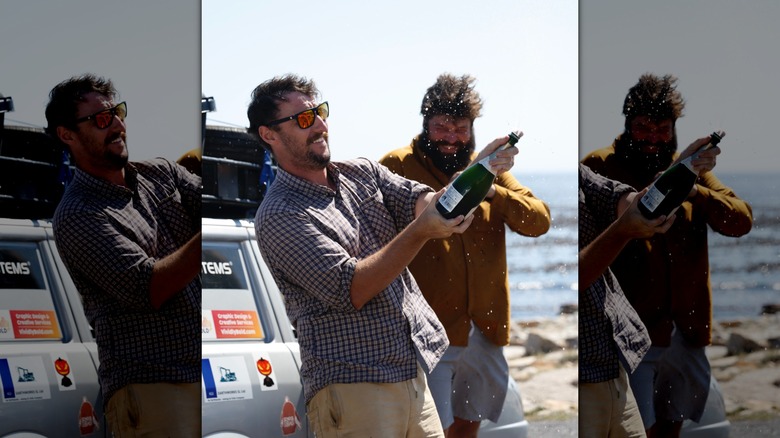 Ollie Jenks pops the cork of a bottle of champagne, with Seth Scott, grinning, standing behind him, and Sheila, the silver overlanding Reliant Robin behind them on a coastal backdrop.