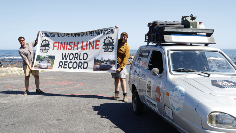 Ollie Jenks (left) and Seth Scott (right) hold a banner behind the silver Robin Reliant called Sheila, marking they reached the finish line for their world record attempt.