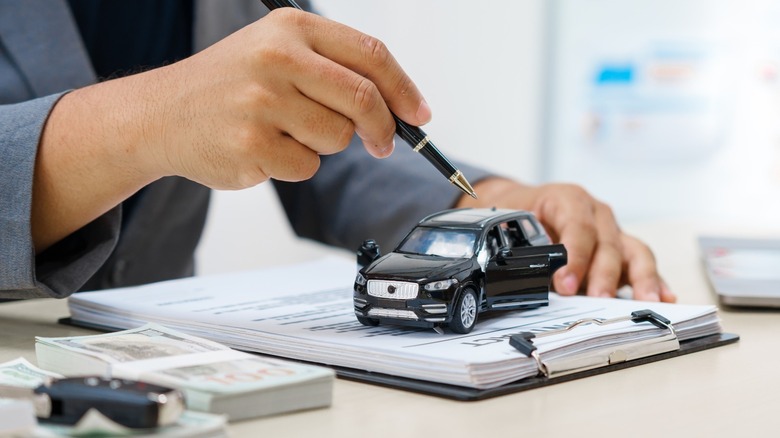 A hand using a pen to point to a toy car on top of a stack of paperwork