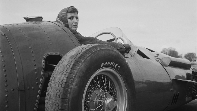 A woman in an old 1950s race car looks over her shoulder from the driver's seat