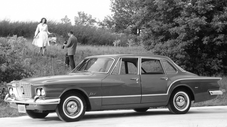 A black and white advertising image of a 1960 Plymouth Valiant with a man and woman in the background. The woman is carrying a picnic basket.