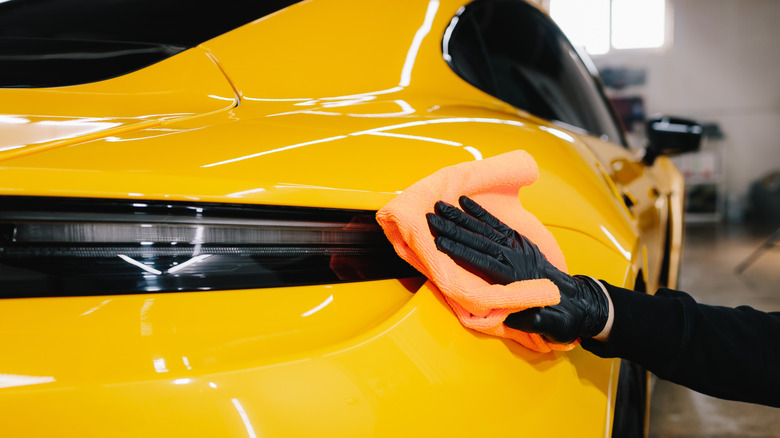 A detailer wearing black gloves wipes a yellow Porsche with a microfiber towel.