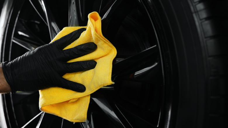 A detailer wearing black gloves wipes a car's wheel with a Kirkland Signature microfiber towel.