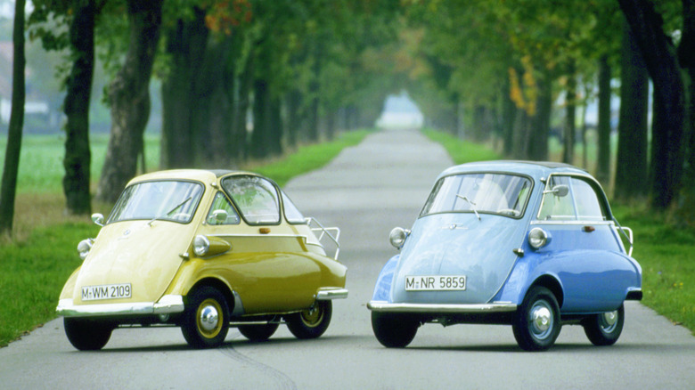 Two BMW Isetta bubble cars, one in yellow/white and one in blue/white, parked on a narrow, tree-lined asphalt road.