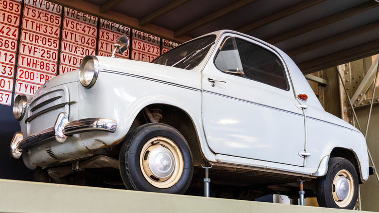 A small white vintage Vespa 400 microcar with a canvas roof, displayed high up on a storage shelf in an indoor museum setting.