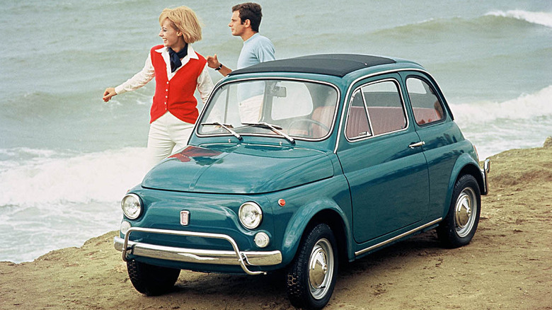 A vintage promotional photograph of a teal-colored 1968-to-1972 Fiat 500 De Luxe parked on a sandy cliffside overlooking the ocean.