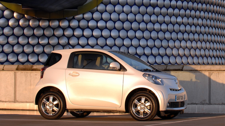 A silver Toyota iQ shown in profile in front of a modern building with a silver, metallic disc-textured facade.