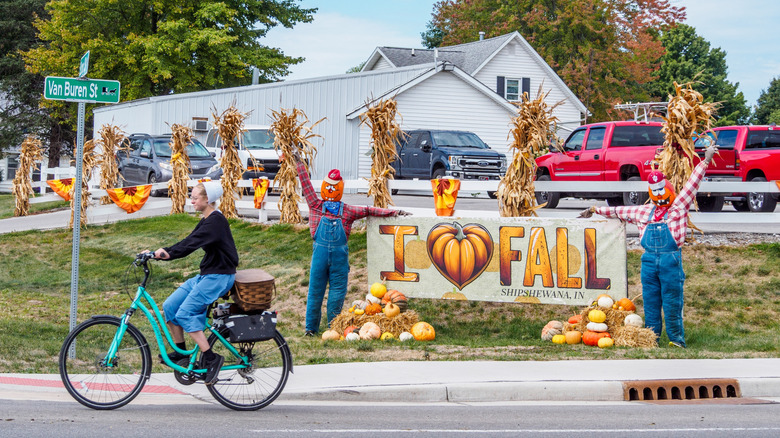 An Amish woman on a bicycle riding by a house with harvest decorations and a "I love fall" sign