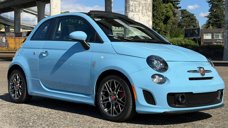 front three-quarters shot of the light blue 500C Abarth parked on asphalt in front of concrete pylons and trees