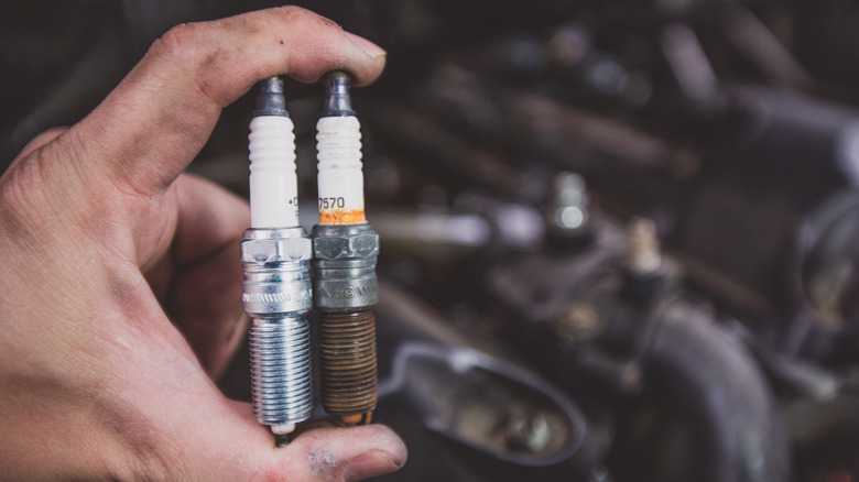 A person holding old and new car spark plugs near an engine