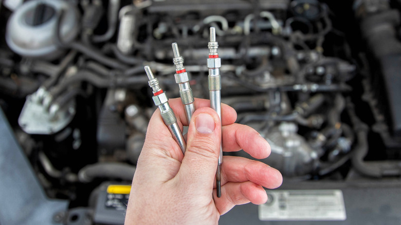 A hand holding three diesel glow plugs with an open hood and engine in background