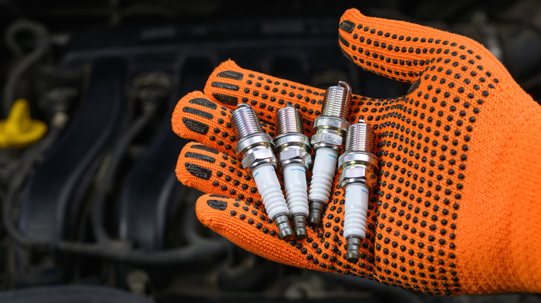 A gloved hand holding spark plugs with an engine in the background.