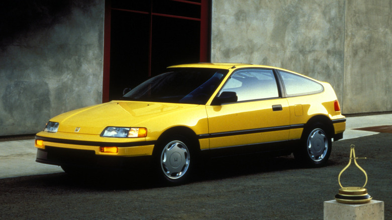 A classic, bright yellow 1988 Honda CRX Si coupe parked against a minimalist concrete wall with a dark, red-framed architectural cutout.