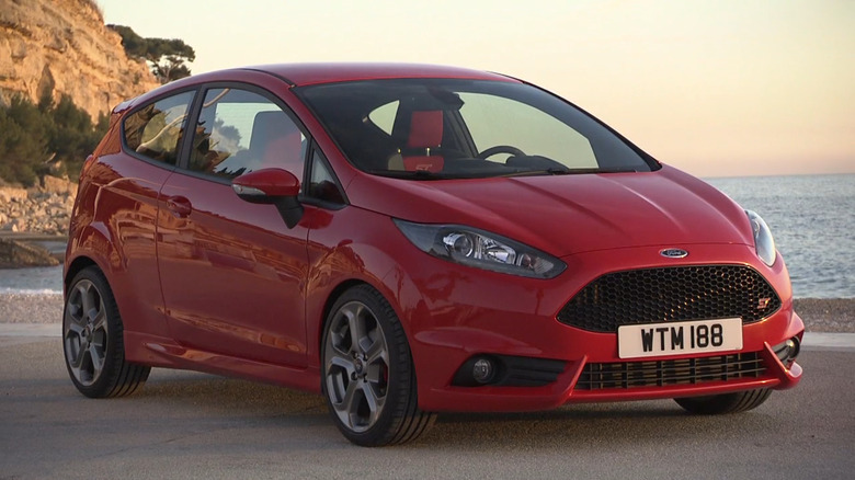 A red Ford Fiesta ST three-door hatchback parked on a coastal overlook at twilight with a calm sea stretching across the background.