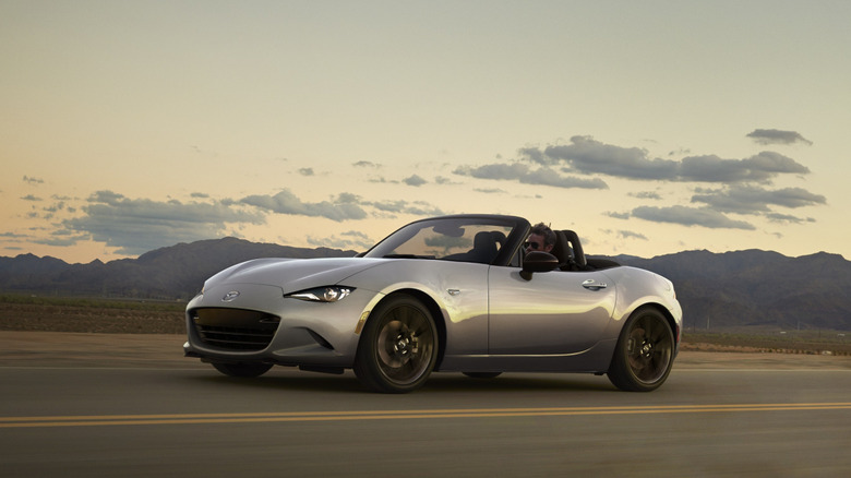 A silver Mazda MX-5 Miata convertible driving down an open desert highway at dusk, with rugged mountain ranges silhouetted against a soft golden hour sky.